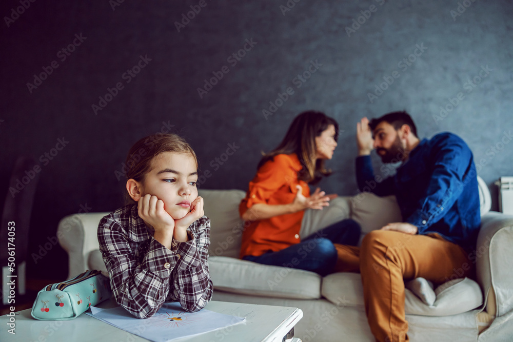 Sad little girl leaning on the desk and listening to parents arguing and yelling.  - obrazy, fototapety, plakaty 