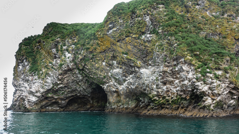 The island in the Pacific Ocean has steep rocky slopes with green vegetation. A dark entrance to the grotto is visible above the water's edge. Kamchatka.