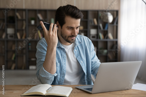 Learning process. Millennial man student prepare for exam at home listen to audio information using smartphone loudspeaker read e book from laptop screen. Young guy work online on pc and mobile ai app