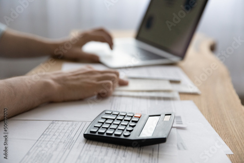 Bookkeeping. Close up of young man accountant financial analyst work with papers invoices statements use computer to declare taxes pay bills on distance. Focus on electronic calculator lying on desk