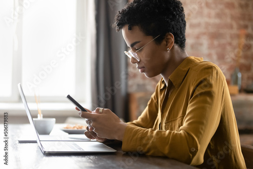 African woman sit at desk holds smartphone. Distracted from work on wireless computer, lead sms chat with client, provide support online, sync information from cellphone to laptop through app concept