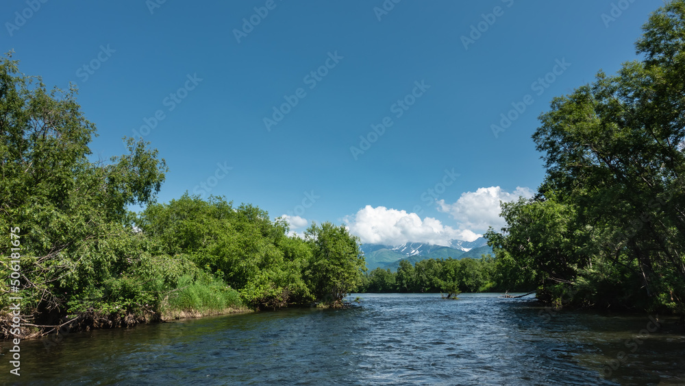 The blue calm river winds between the banks overgrown with lush green vegetation. In the distance, a mountain range is visible, hiding in the clouds. Kamchatka The blue calm river winds between the banks overgrown with lush green vegetation. In the distance, a mountain range is visible, hiding in the clouds. Kamchatka