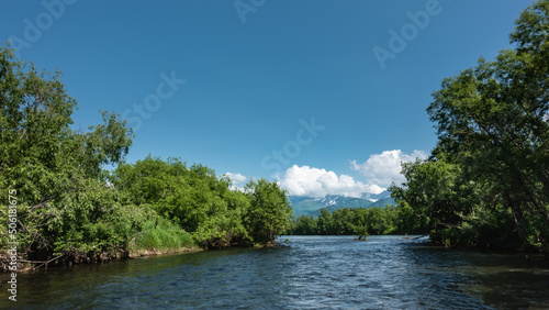 The blue calm river winds between the banks overgrown with lush green vegetation. In the distance, a mountain range is visible, hiding in the clouds. Kamchatka