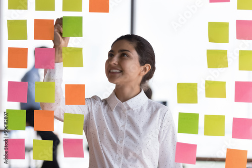Fotografiet Attractive young Indian female employee jotting information and reminders on colorful sticky notes attached on glass wall in office