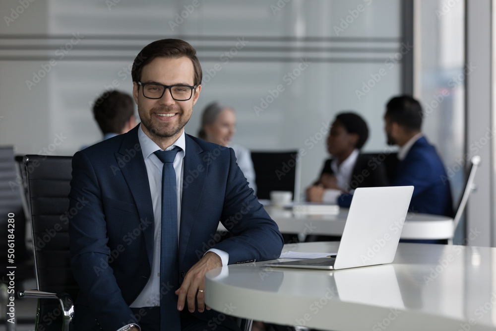 Handsome millennial smiling businessman in elegant formal suit sit at desk with laptop staring at cam, diverse businesspeople on background. Leadership, careerism, business application usage concept Handsome millennial smiling businessman in elegant formal suit sit at desk with laptop staring at cam, diverse businesspeople on background. Leadership, careerism, business application usage concept