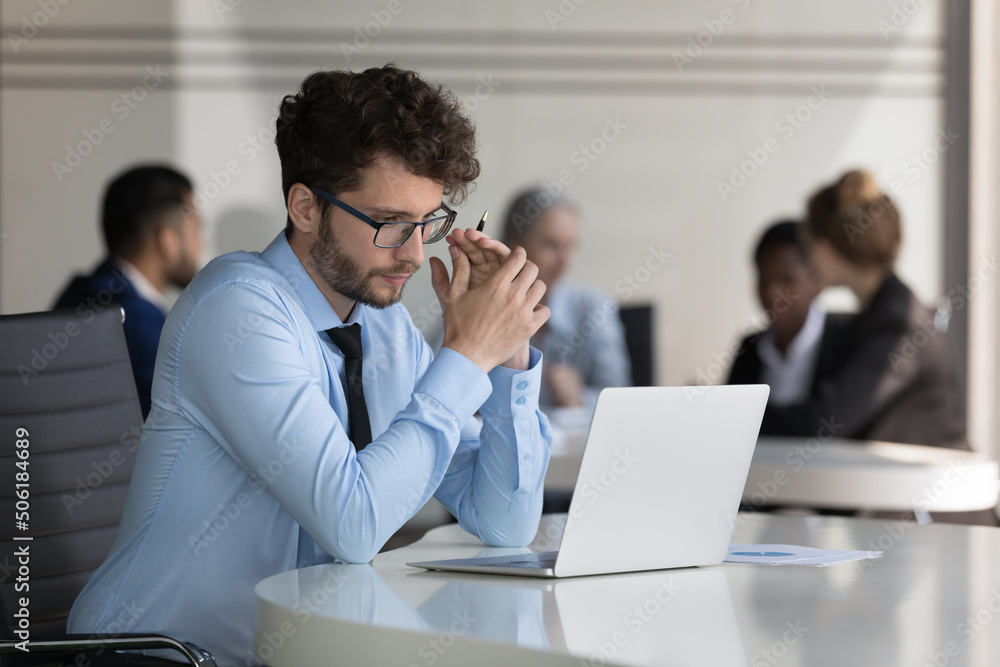 Young serious pensive businessman work on market research sit at desk with laptop in modern office with diverse businesspeople on background. Modern tech, business project, challenge, workflow concept Young serious pensive businessman work on market research sit at desk with laptop in modern office with diverse businesspeople on background. Modern tech, business project, challenge, workflow concept
