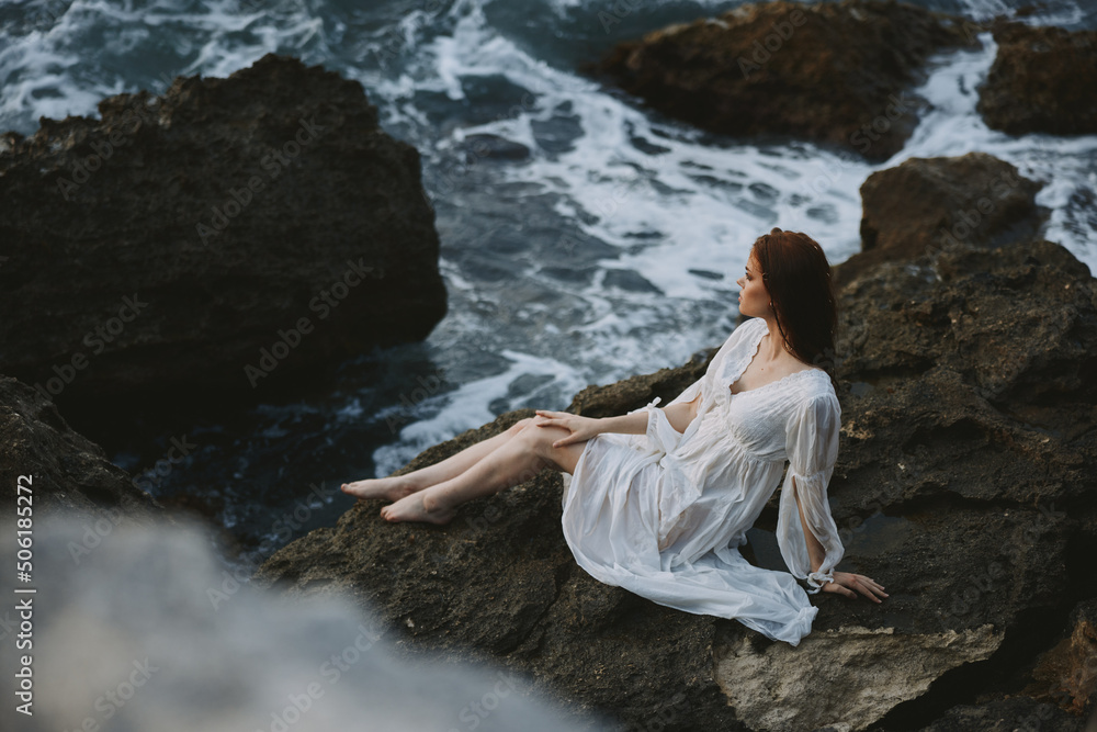 A woman in a white wedding dress sits on a rocky stone by the ocean just