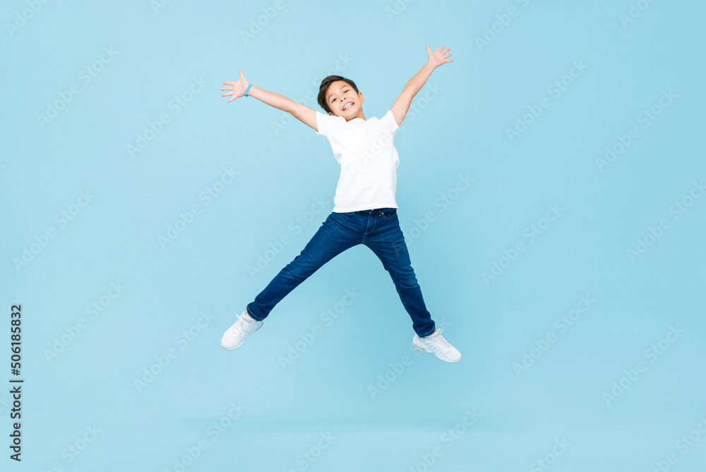 Energetic Asian mixed race boy jumping and raising hands up in isolated light blue color studio background