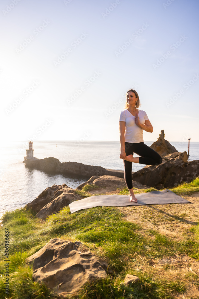 A blonde woman doing yoga exercises in nature by the sea, stretching next to a lighthouse, healthy and naturist life, pilates outdoors