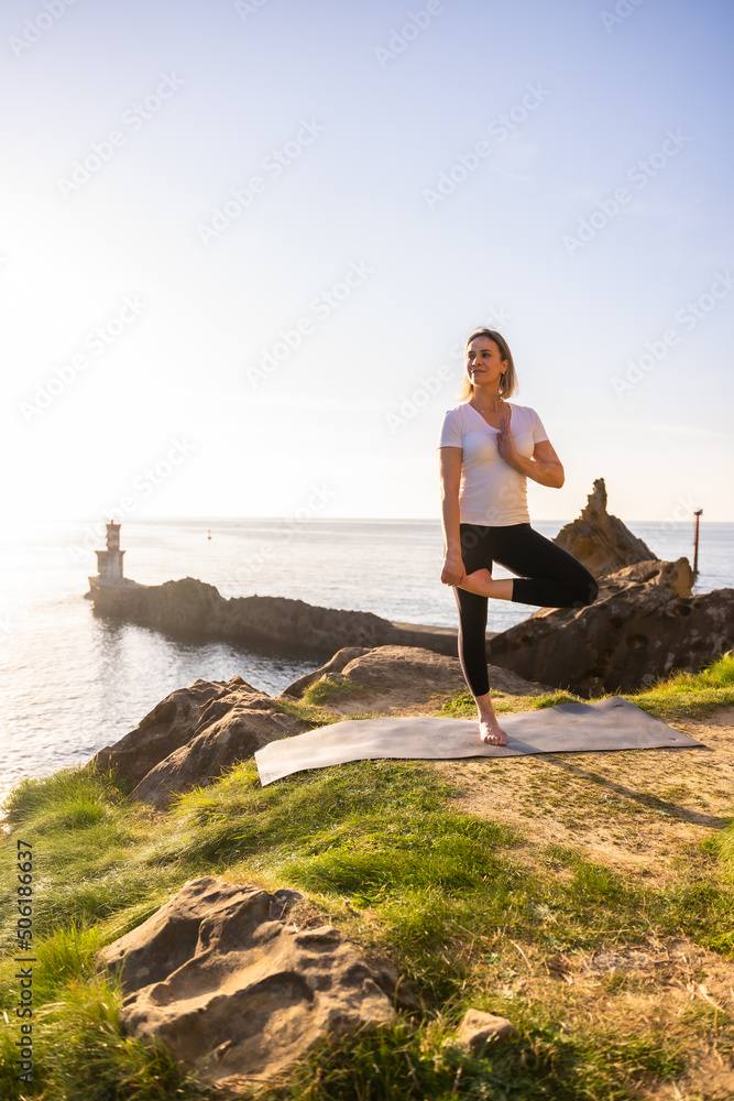 A blonde woman doing yoga exercises in nature by the sea, stretching next to a lighthouse, healthy and naturist life, pilates outdoors A blonde woman doing yoga exercises in nature by the sea, stretching next to a lighthouse, healthy and naturist life, pilates outdoors