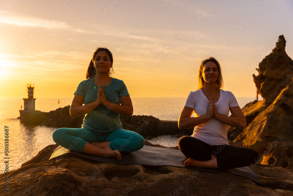 Two women sitting on yoga mat in nature by the sea at sunset, healthy and naturist lifestyle, outdoor pilates, ardha padmasana