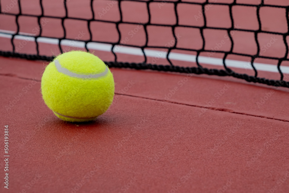 Tennis ball next to the net of a red hard surface tennis court.