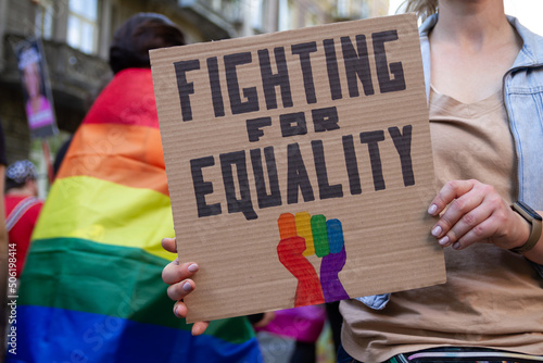 Woman holding placard sign Fighting for Equality with rainbow flag fist, during LGBT Pride Parade. Crowd of people at equality march to support and celebrate LGBT+, LGBTQ Gay and lesbian community.