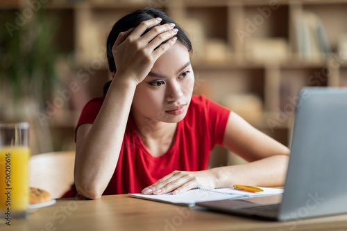 Tired unhappy pretty young asian woman student looking at laptop in living room interior