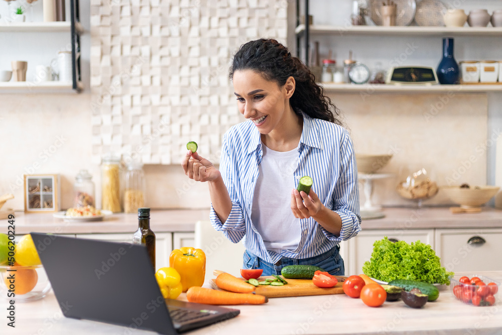 Happy latin woman cooking dinner and video calling on laptop, talking to webcamera, standing in kitchen at her apartment Happy latin woman cooking dinner and video calling on laptop, talking to webcamera, standing in kitchen at her apartment