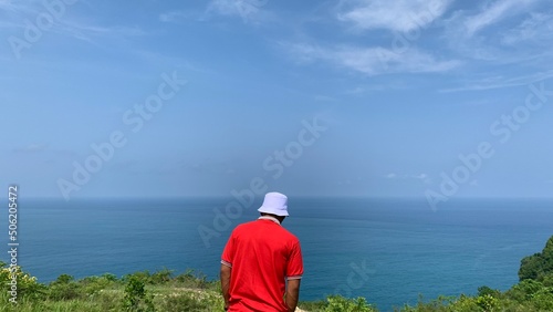 a man in a red shirt and a white bucket hat standing and walking on a hill looking at the beautiful view of the open sea