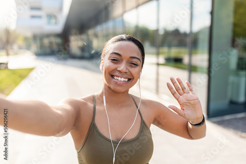 Happy black female blogger taking selfie or greeting followers, waving at webcam on mobile phone, jogging outdoors © Prostock-studio