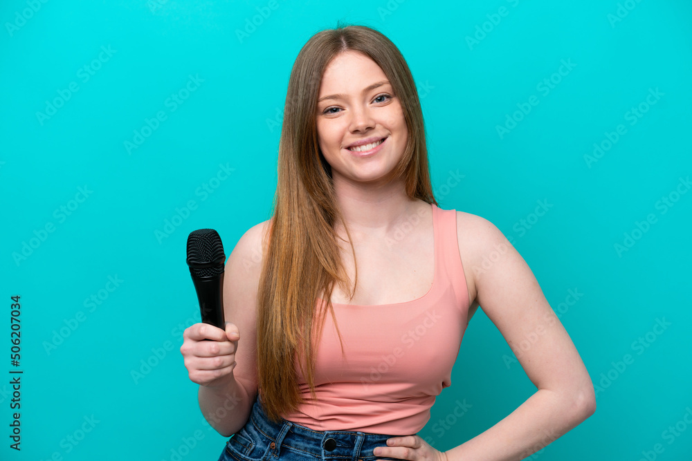 Singer caucasian woman picking up a microphone isolated on blue background posing with arms at hip and smiling Singer caucasian woman picking up a microphone isolated on blue background posing with arms at hip and smiling