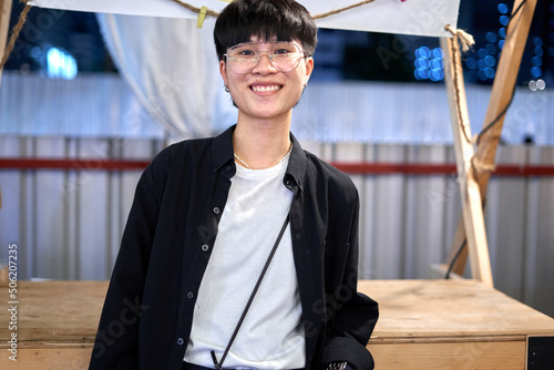 Smiling non-binary person with glasses standing in an urban night fair