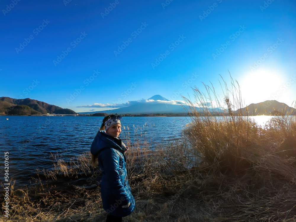 A woman standing in between golden grass at the shore of Kawaguchiko Lake, Japan with the view on Mt Fuji. The mountain surrounded by clouds. Soft reflections in the calm surface of the lake. Serenity