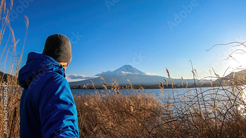 Man walking through golden grass at the side of Kawaguchiko Lake, Japan with the view on Mt Fuji, while taking a selfie. The mountain surrounded by clouds. Serenity and calmness. Exploring new places