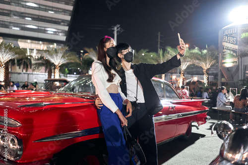 Lesbian couple taking a selfie while embracing at a night fair with antique cars