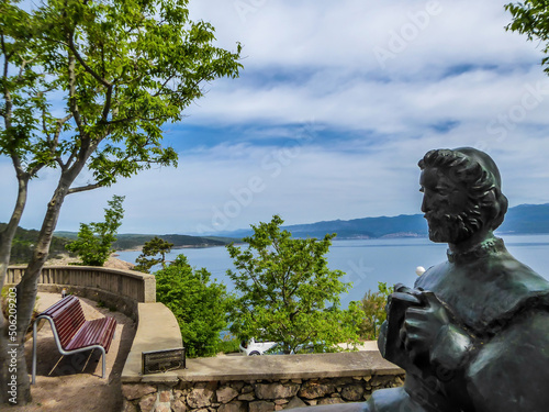 Vrbnik/Croatia-05/01/2018: A statue of a man, located at the top of a hill with a beautiful view on the sea. Some trees disturbing the view on the sea. Bench vith the sea view.