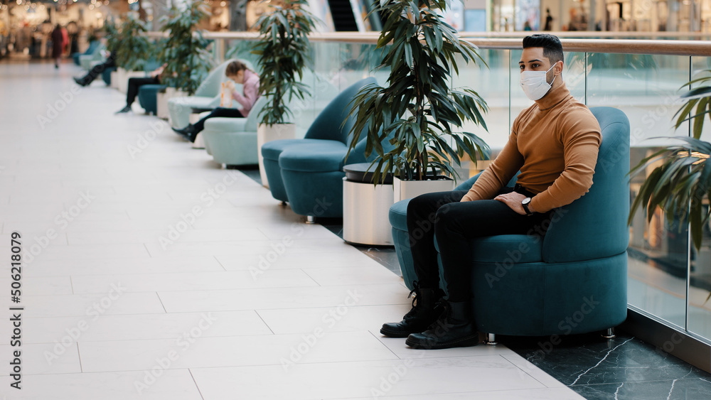 Obraz premium Young indian guy in medical mask protecting health during pandemic sitting in armchair indoors in shopping center mall resting relaxing nervous anxious waiting for appointment long expectation concept