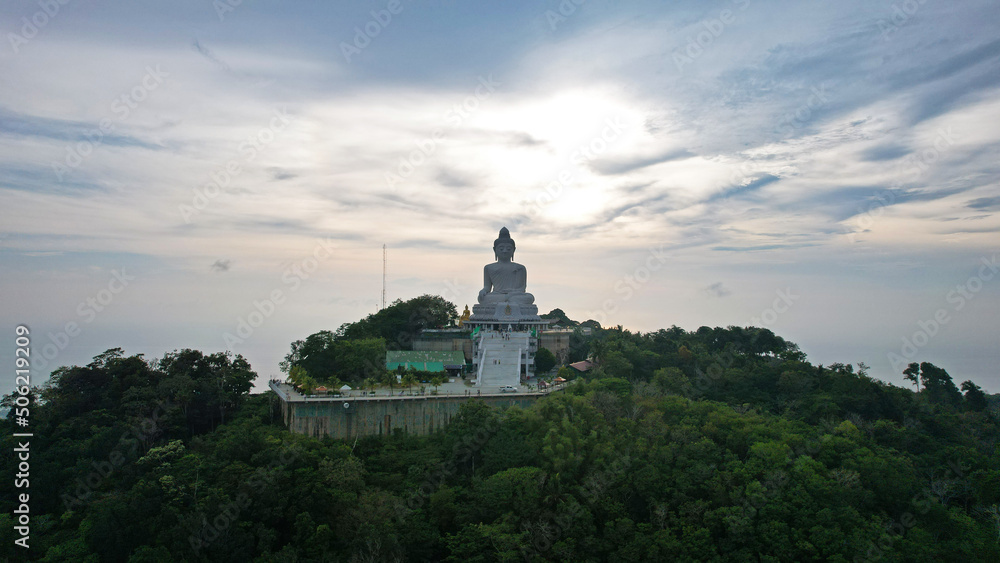 Drone view of the Big Buddha, Thailand. The Big Buddha is sitting on hill in the lotus position, meditating. The sun shines brightly through clouds. There's a jungle all around. A ray of sun on water