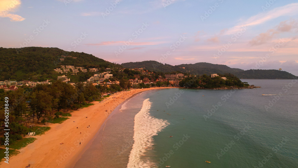 Orange sunset with a view of the beach. People are resting, seeing off the sun over the horizon. Small waves. Clean sand. Hills, clouds and the city are visible in the distance. Dogs are running. Kata