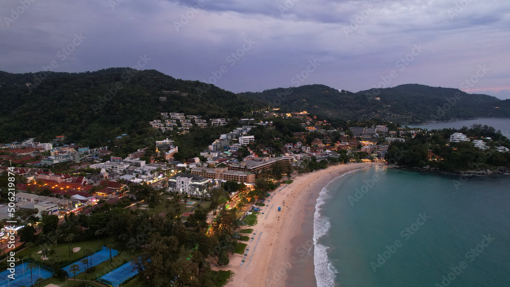 Aerial view of the island at sunset from a drone. Tall palm trees and trees grow. In the distance there are hotels, sports fields, green hills and pink clouds. A place to rest. Phuket Island, Kata