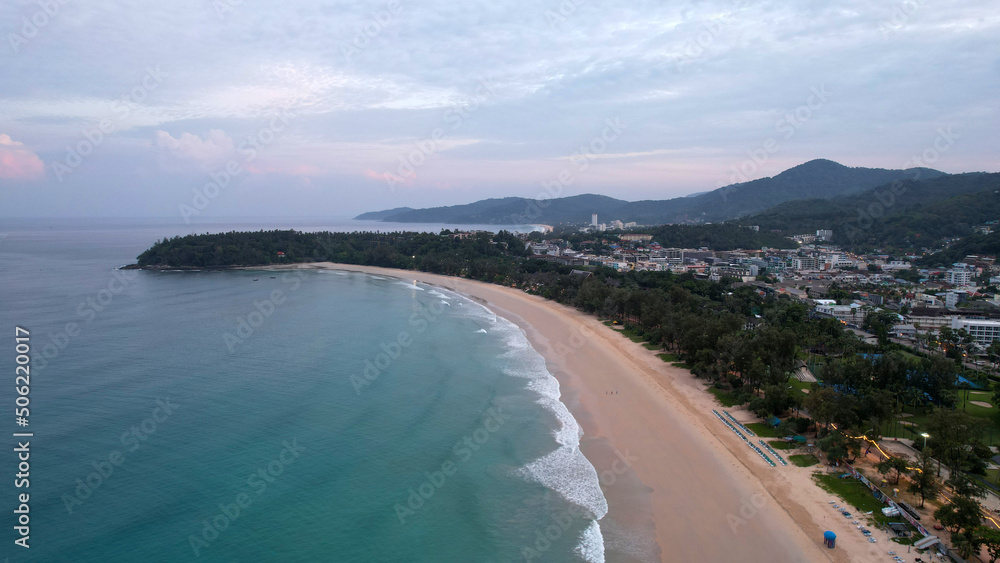 Early dawn on the island of Phuket. Top view from the drone on the sand and waves. People are walking along the beach. Calm environment. Green hills and clouds in sky. Lights of the city are shining