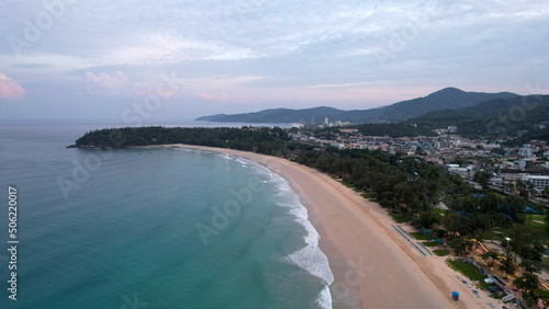 Early dawn on the island of Phuket. Top view from the drone on the sand and waves. People are walking along the beach. Calm environment. Green hills and clouds in sky. Lights of the city are shining