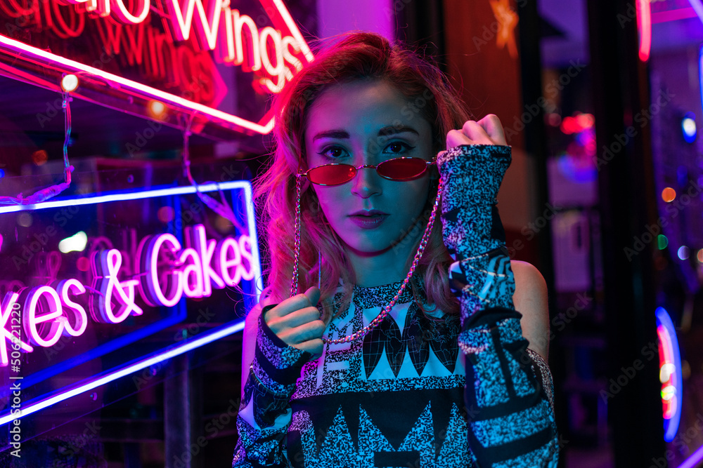 Millennial enigmatic pretty girl with unusual dyed hairstyle near glowing neon wall at night. Blue hair, golden sequins as freckles,nose piercing. Mysterious hipster teenager in glasses - obrazy, fototapety, plakaty 