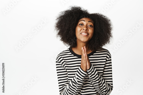 Portrait of Black girl asking for help, say please, begging, yearning to achieve smth, holding hands in pleading gesture, standing over white background © Cookie Studio