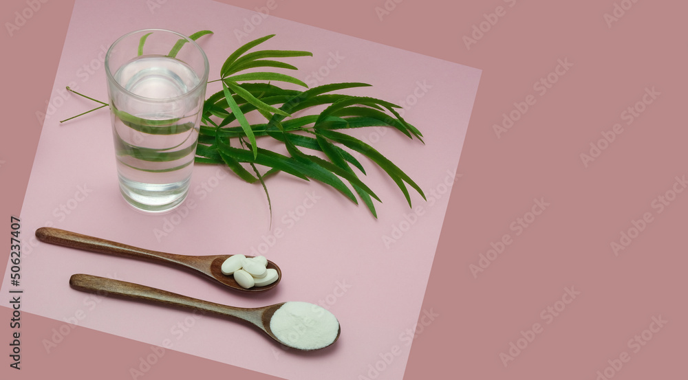 Collagen powder and tablets in wooden spoons on a combined pink background. A clear glass of water and beautiful green leaves. Horizontal banner with copyspace.