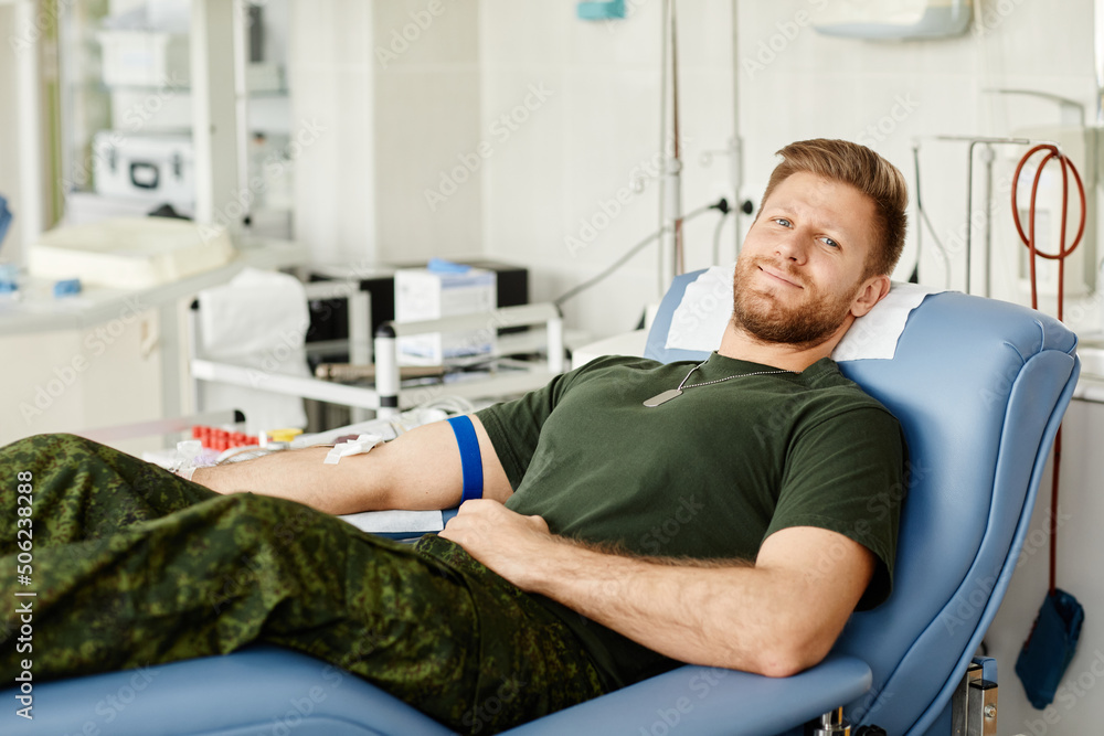Obraz premium Portrait of military man giving blood while laying in chair at blood donation center and smiling at camera, copy space