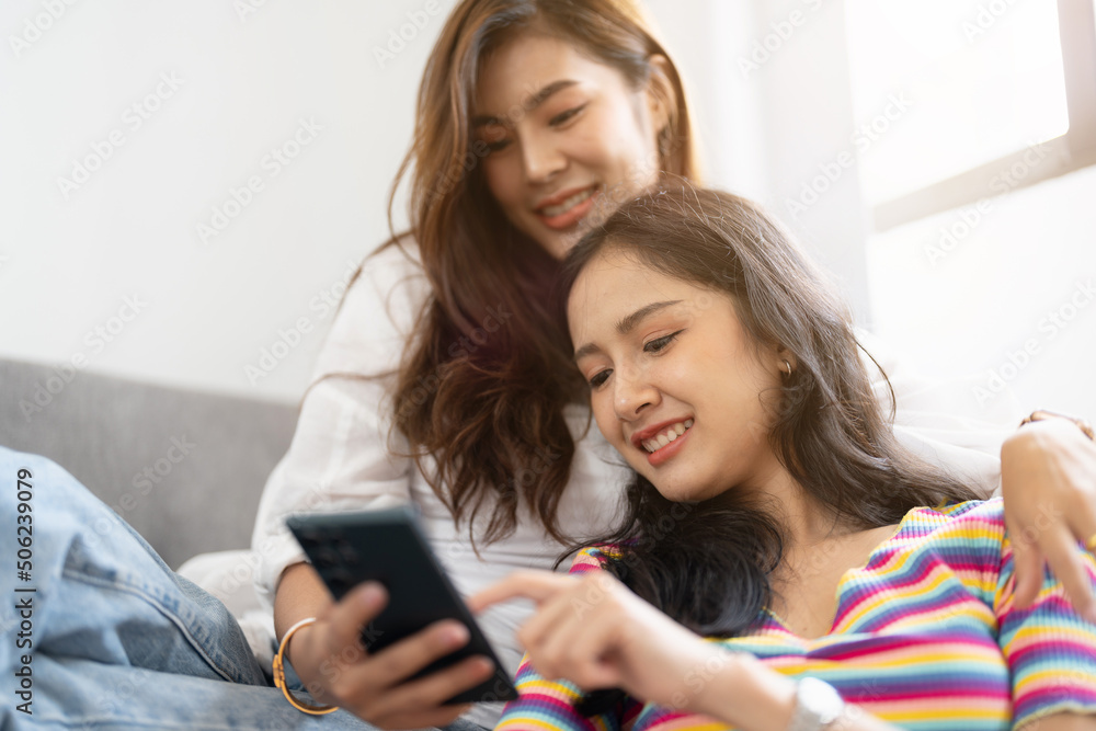 Two happy young aisan casual women having fun using smartphone on couch at home. Lesbian millennial couple. gbt, homosexual, lesbian couple lifestyle.
