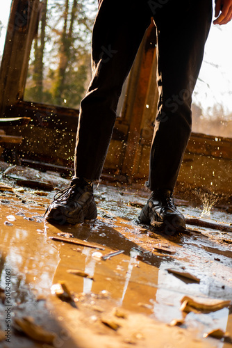 Male legs in black pants and sneakers among the puddle and frozen drops in a flooded, destroyed and abandoned house. Solar rays. Consequences of a natural disaster. Vertical shot.