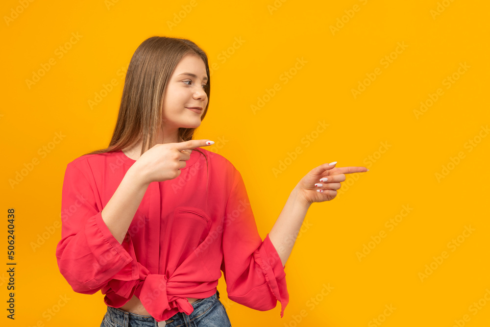 Beautiful girl teenager smiling and pointing finger aside isolated over orange background. Copy space. Beautiful girl teenager smiling and pointing finger aside isolated over orange background. Copy space.