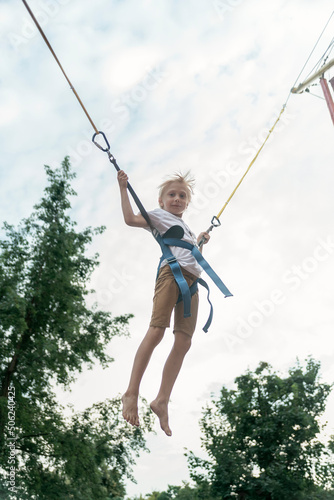 Blond boy jumping in an amusement park on the ropes high into sky. Child has fun in the theme park.
