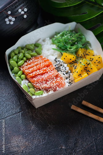 Poke with salmon, mango, edamame and seaweed in a carton delivery container, vertical shot on a dark-brown stone surface, middle closeup