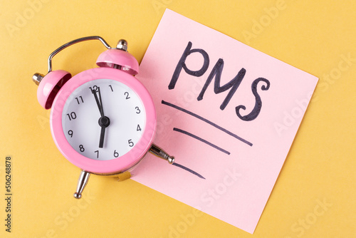 Pink alarm clock (clock), the inscription PMS on a piece of paper on a yellow background. Women's health, menstruation. The concept of the critical days period.