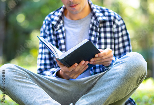 Young student wearing a plaid shirt sitting on the grass reading a book