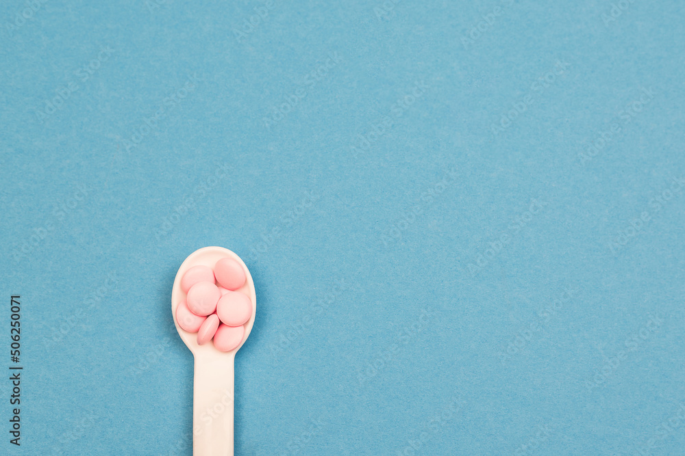 A white spoon with pink pills on a blue background. The concept of health and pharmaceuticals. Copy space. A white spoon with pink pills on a blue background. The concept of health and pharmaceuticals. Copy space.