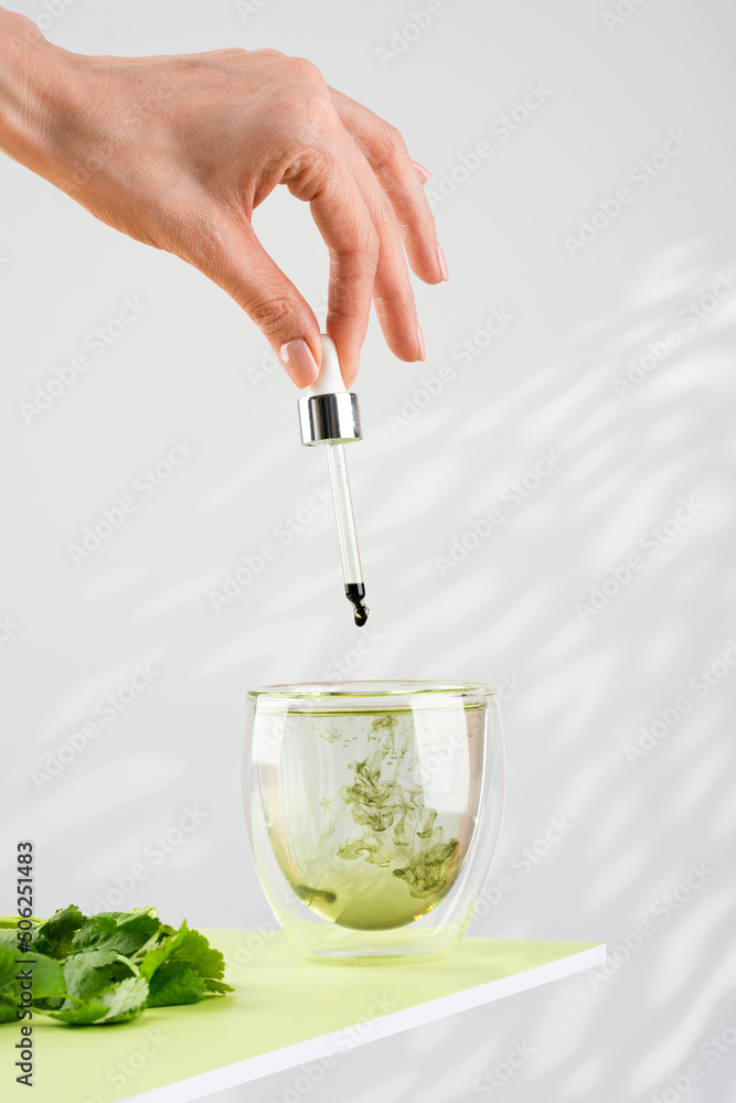 Female hand pours liquid chlorophyll into a glass of water with a dropper. Glass of liquid chlorophyl and fresh herbs on the green table. Gray background, natural light. Concept of superfood, healthy - obrazy, fototapety, plakaty 