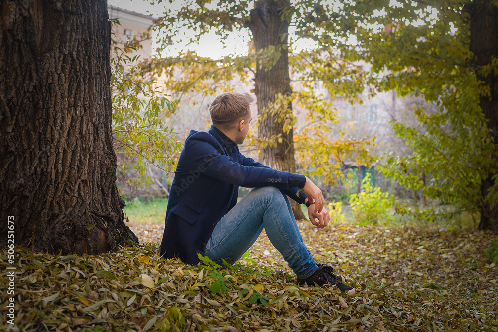 Wistful man in the suit sits on the ground in the autumn foliage in the park.