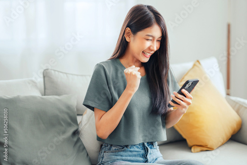 Excited young asian woman hold phone chatting in dating app feel happy sit on sofa at home, Closeup joyful female reading good news on phone. Surprised lady celebrating victory on phone photo