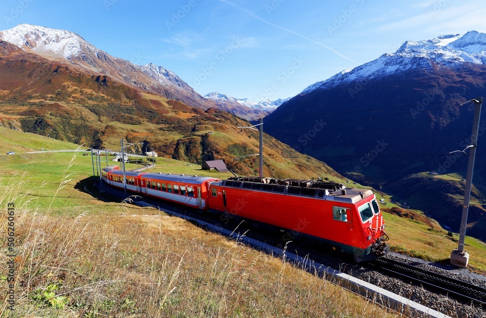 A tourist train traveling on the green grassy hillside on a beautiful autumn day and snowy alpine mountains towering under blue sunny sky in background, in Andermatt, canton of Uri, Switzerland
