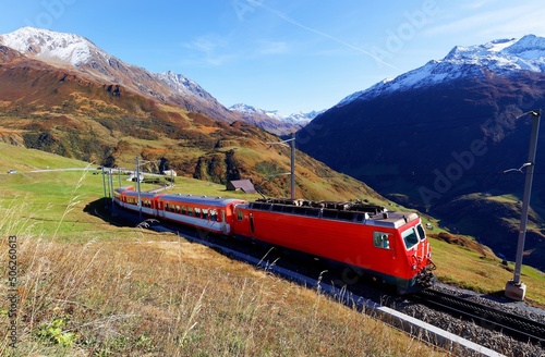 A tourist train traveling on the green grassy hillside on a beautiful autumn day and snowy alpine mountains towering under blue sunny sky in background, in Andermatt, canton of Uri, Switzerland